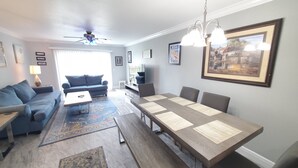 Ein modernes Wohnzimmer mit Sofa, Couchtisch und Esstisch. Es gibt einen Deckenventilator, einen Fernseher und gerahmte Bilder an der Wand.