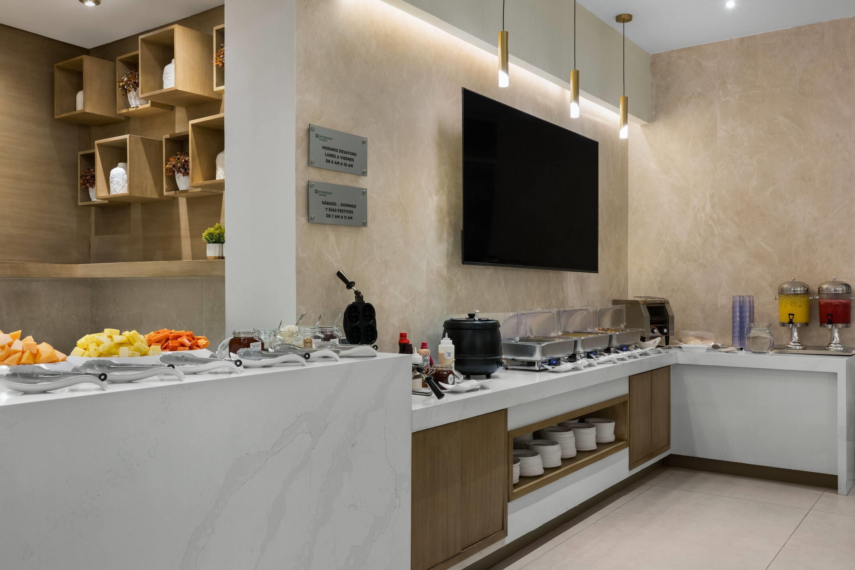 A breakfast bar with various food items and condiments, a flat-screen TV mounted on the wall, and a sign indicating the breakfast hours.