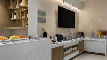 A breakfast bar with various food items and condiments, a flat-screen TV mounted on the wall, and a sign indicating the breakfast hours.