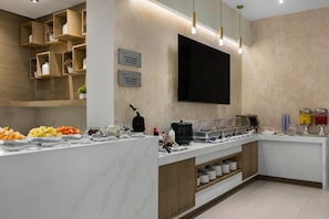 A breakfast bar with various food items and condiments, a flat-screen TV mounted on the wall, and a sign indicating the breakfast hours. - Wyndham Garden Saltillo (Saltillo)