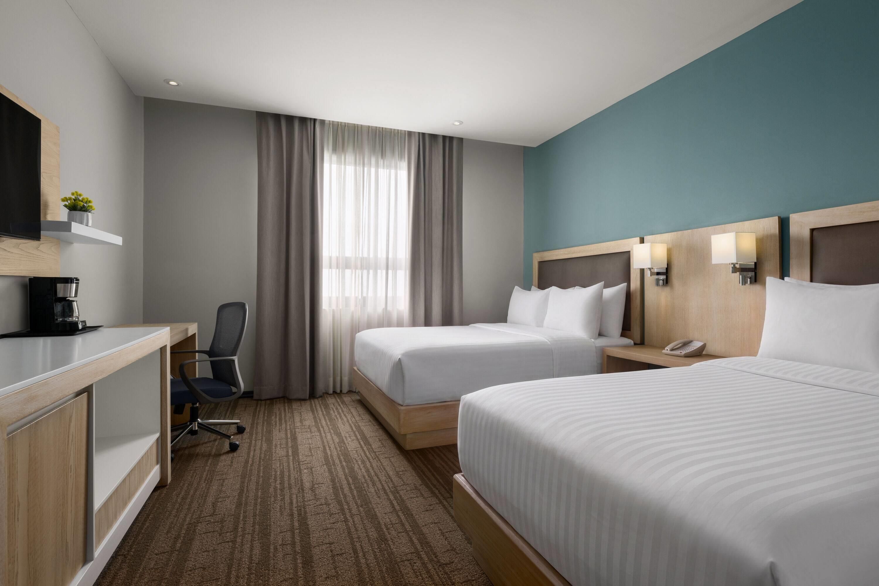 Habitación de hotel con dos camas, un escritorio con cafetera, una silla y un ventanal con cortinas.