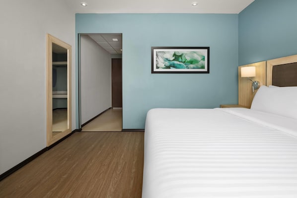 A hotel room with a bed, a nightstand, a wall-mounted lamp, a framed abstract painting, and a closet with a mirror.