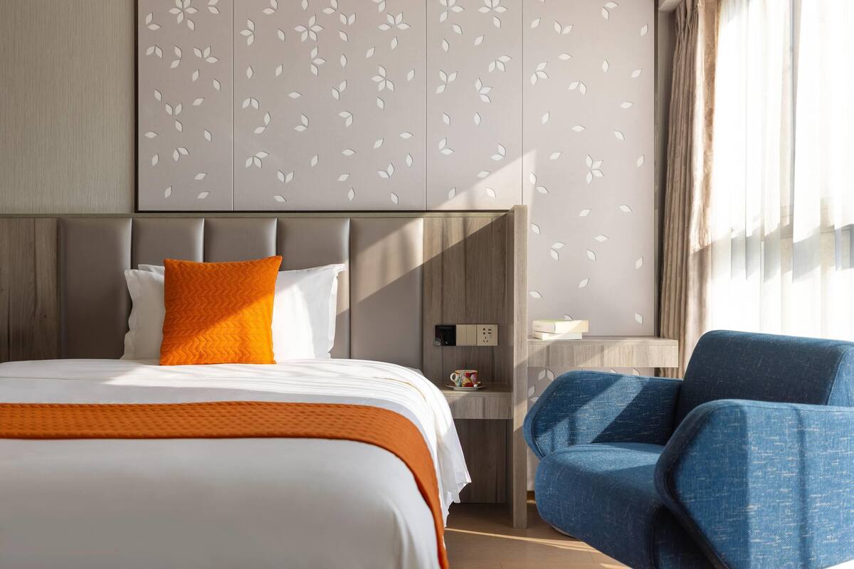 Hotelkamer met een bed, een oranje plaid, een blauwe fauteuil, een nachtkastje met een lamp, en een muur met een bladmotief.