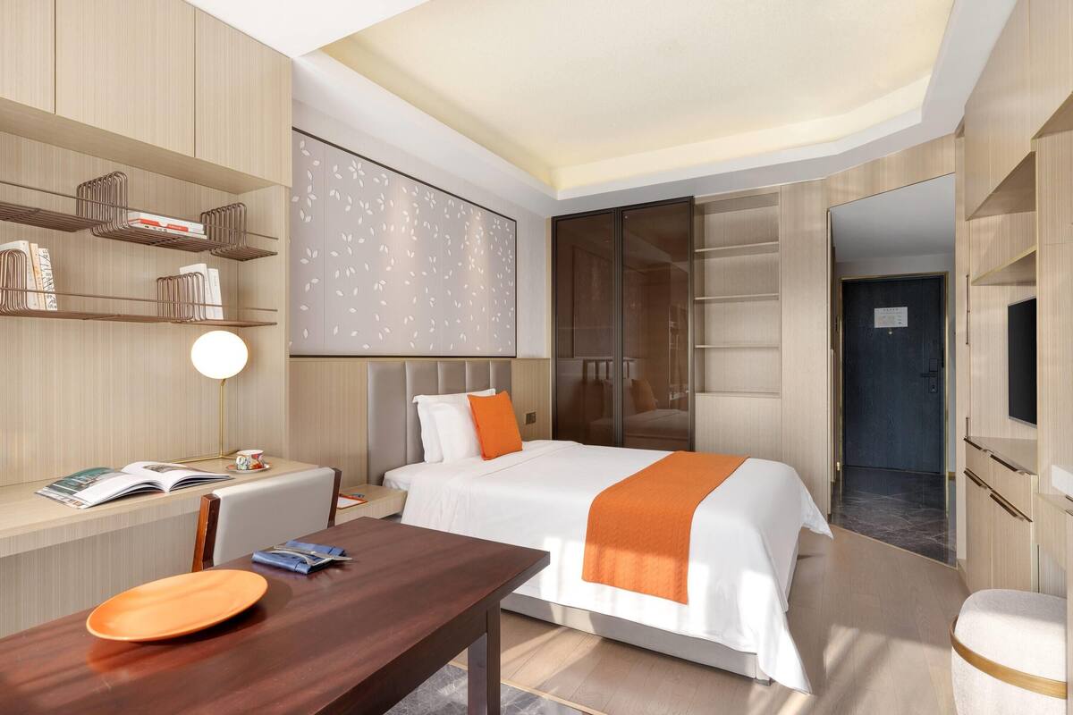 Een moderne hotelkamer met een opgemaakt bed, een bureau met lamp en ingebouwde planken met boeken.
