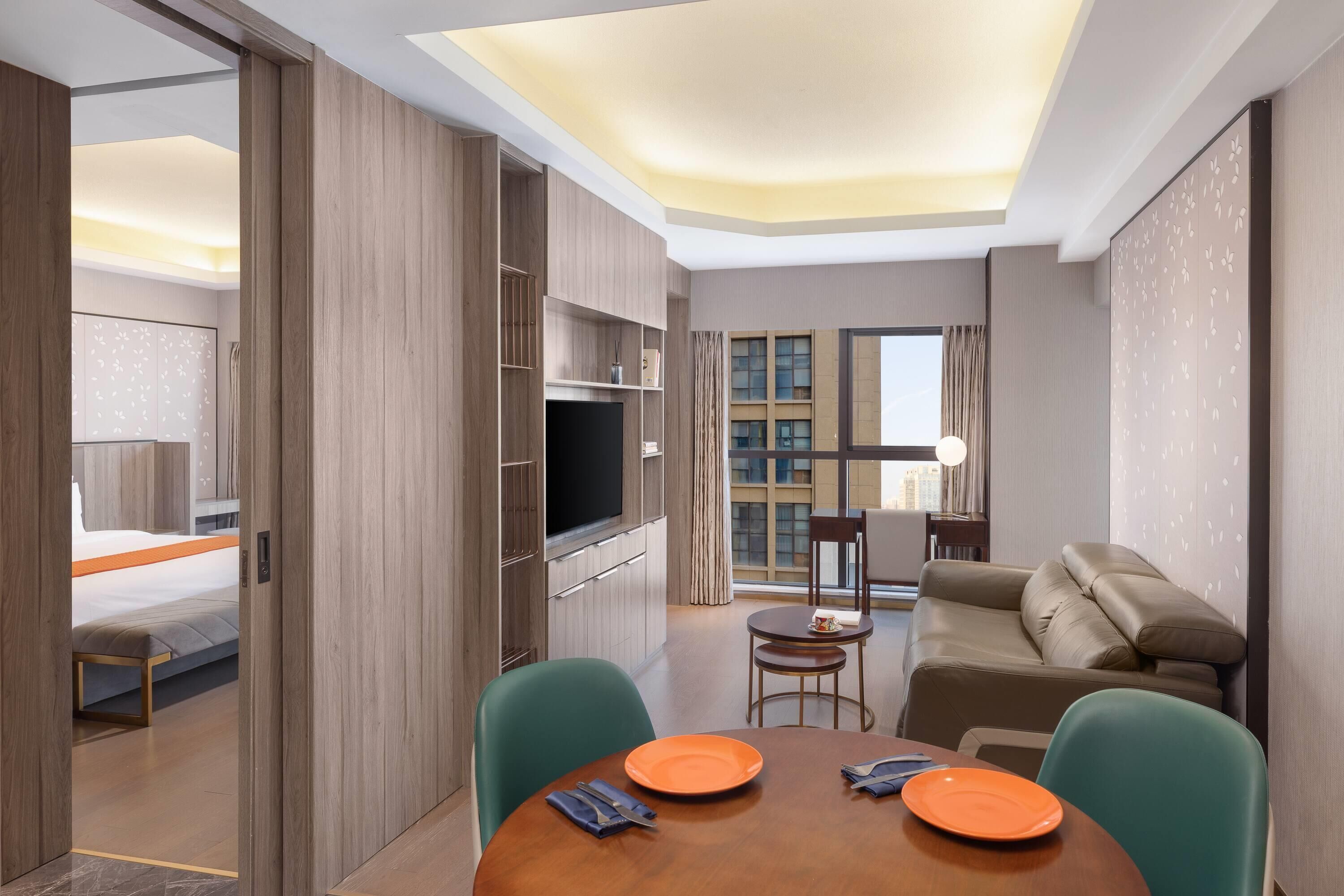 A modern hotel room with a wooden wardrobe, a flat-screen TV, a sofa, a dining table with plates, and a large window offering a city view.
