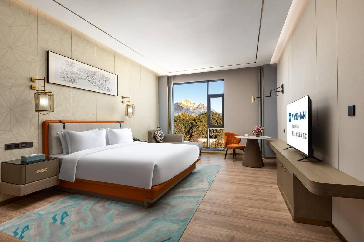 A modern hotel room with a bed, bedside tables, a desk, a TV, and a large window with a mountain view.