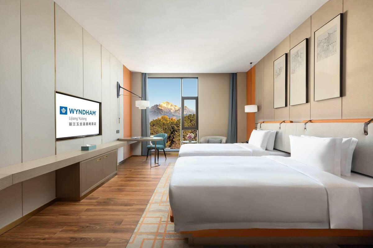 A hotel room with a bed, a desk, a chair, and a large window with a mountain view.