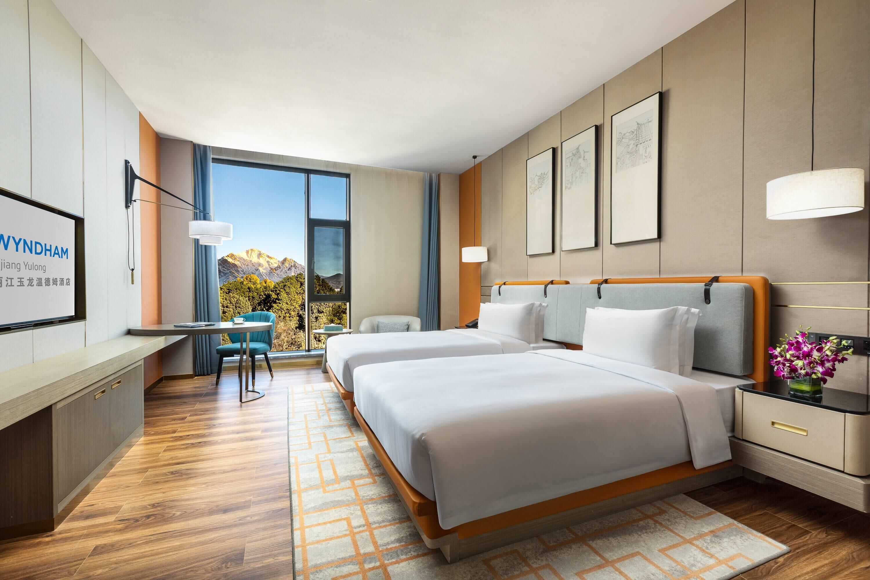 A modern hotel room with two beds, a desk, a chair, and a large window with a mountain view.