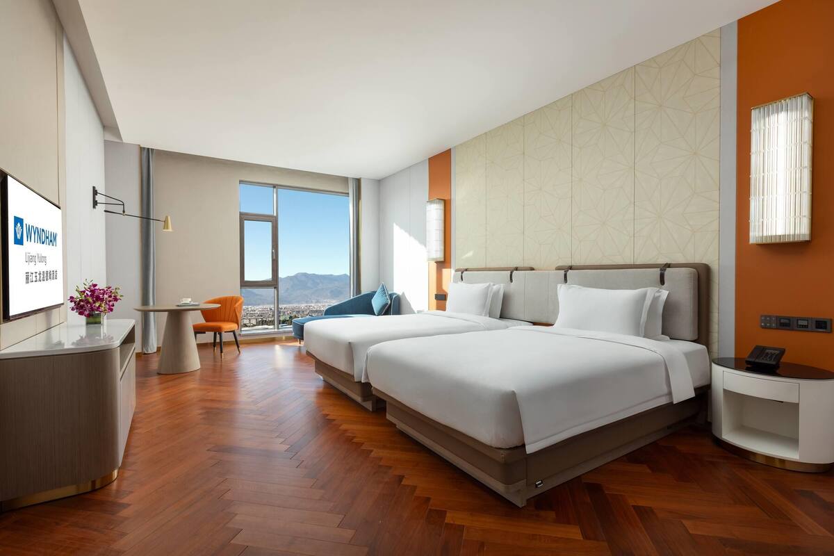 A hotel room with two beds, a desk, a chair, a small table, and a large window offering a city view.