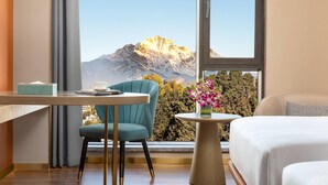 Een hotelkamer met een groot raam, een bed, een ronde tafel met een bloevas, een stoel en een klein tafeltje met een kopje en zakdoekjes.
