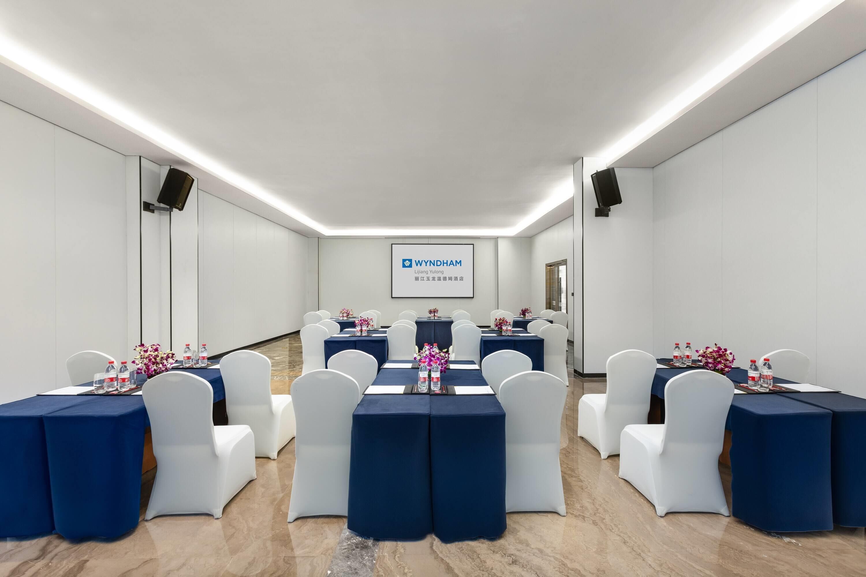 A conference room with white chairs and tables, a projector screen, and a logo on the wall.