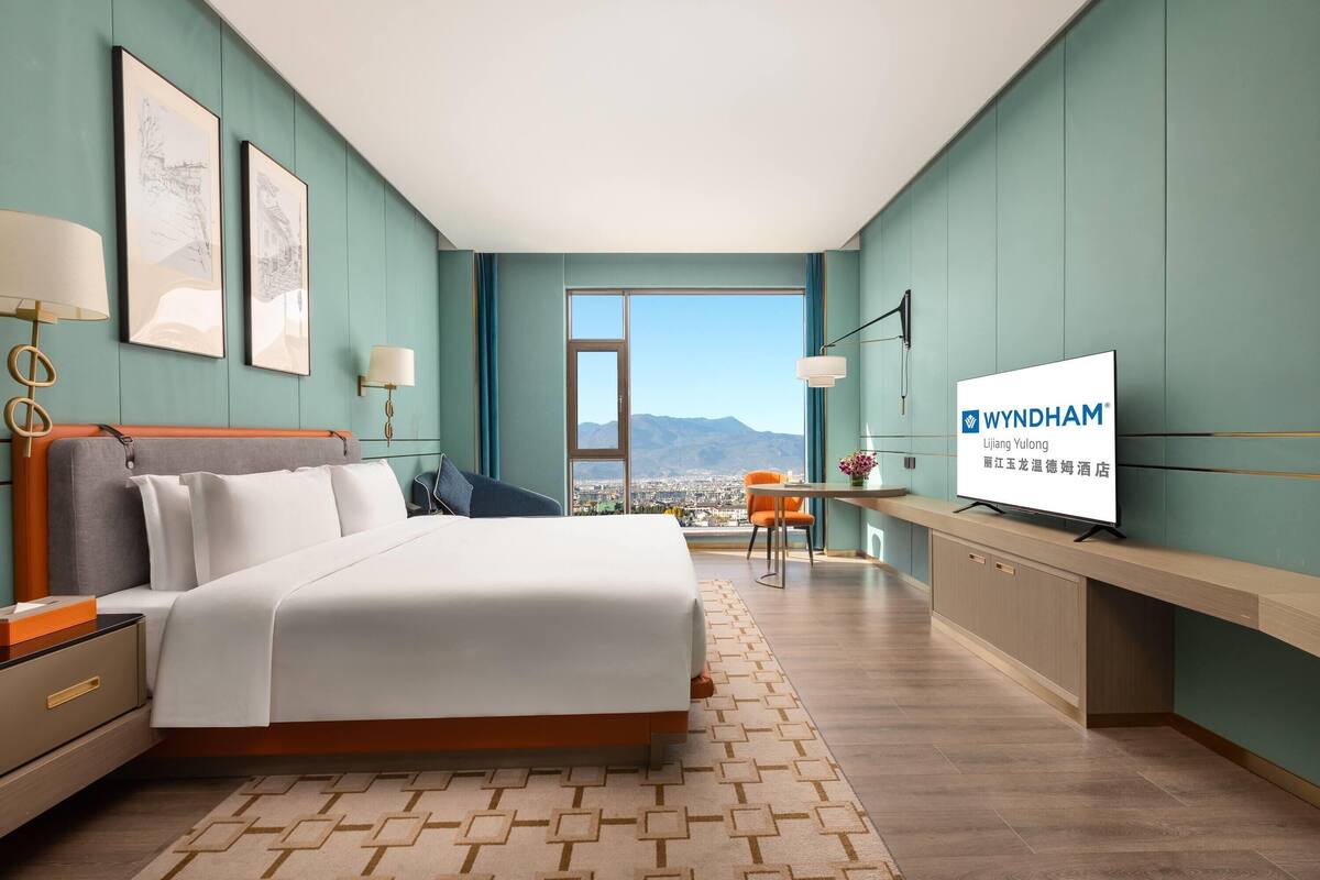A hotel room with a bed, bedside table, lamp, artwork, TV, and a view of the city and mountains.