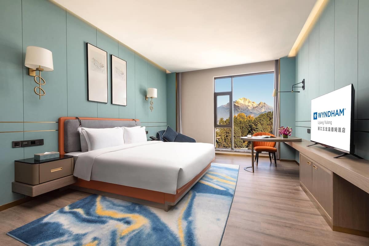 A modern hotel room with a bed, bedside tables, a desk with a chair, a flat-screen TV, and a large window with a mountain view.