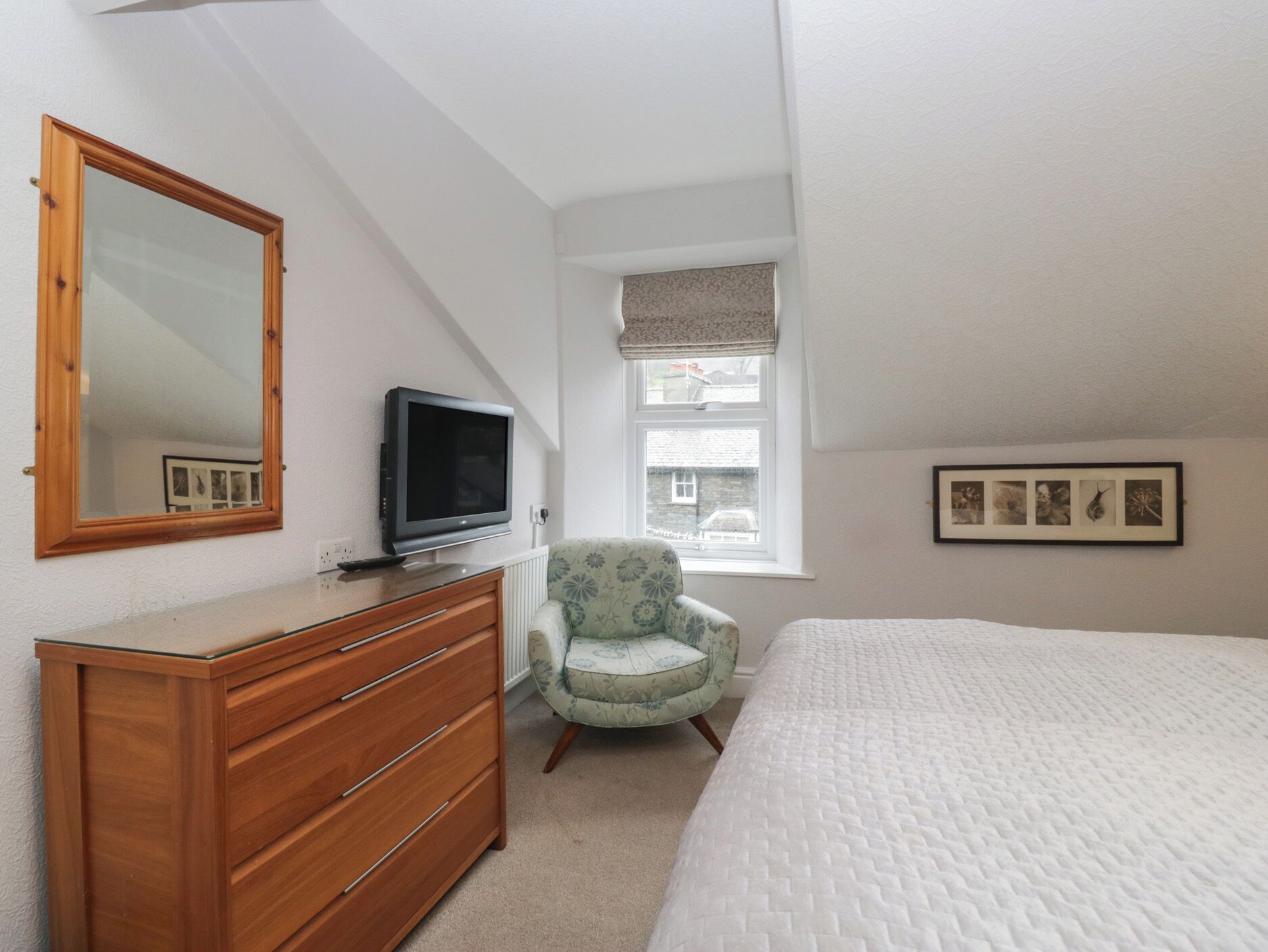 A bedroom with a wooden dresser, a television, a floral armchair, a window with a view, and a framed photo on the wall.