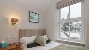A bedroom with a bed, a bedside table, a lamp, a framed picture, and a window with a view of buildings. - Norwood House (Ambleside)