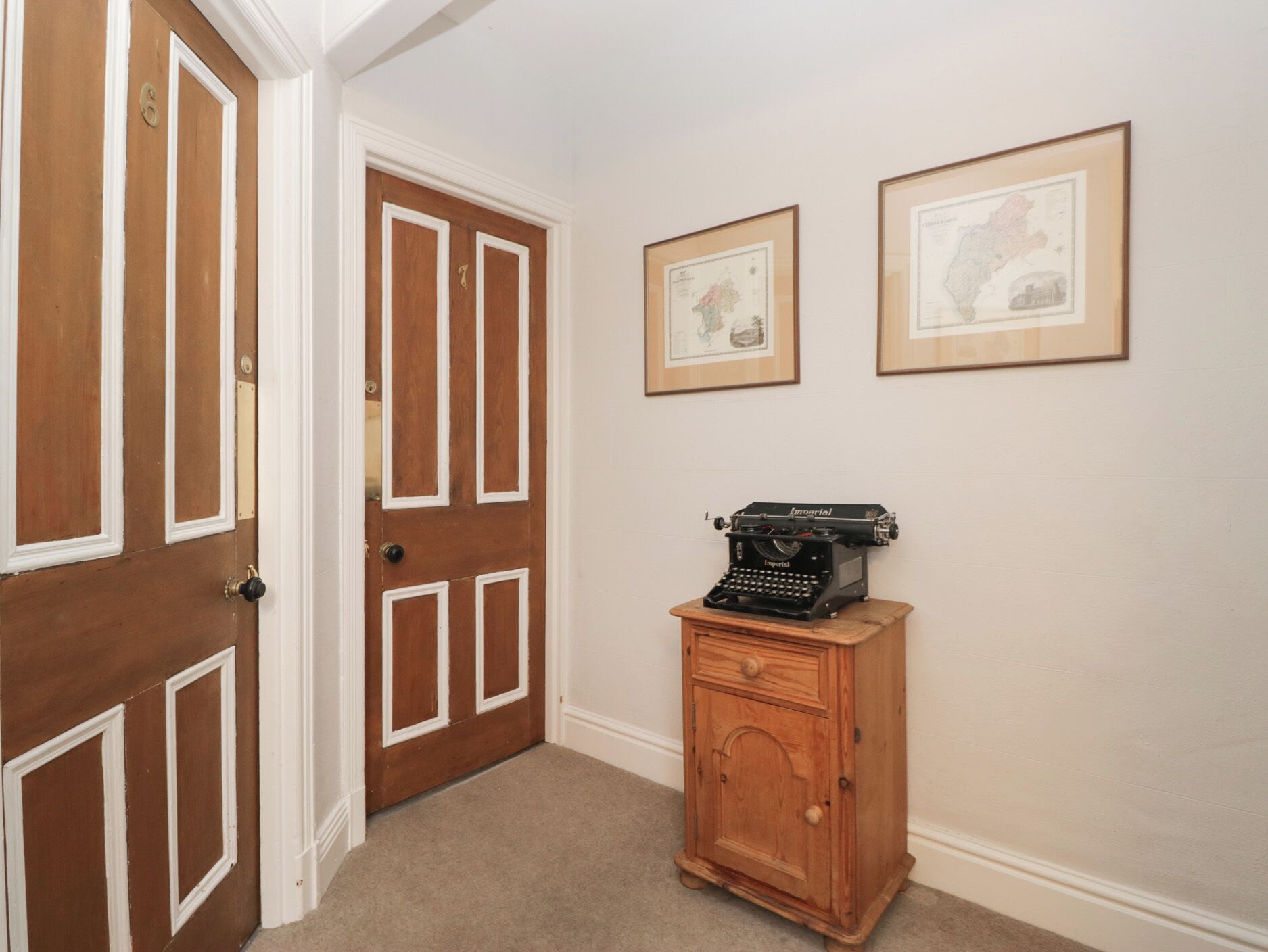 A hallway with wooden doors, a vintage typewriter on a wooden cabinet, and two framed maps on the wall.