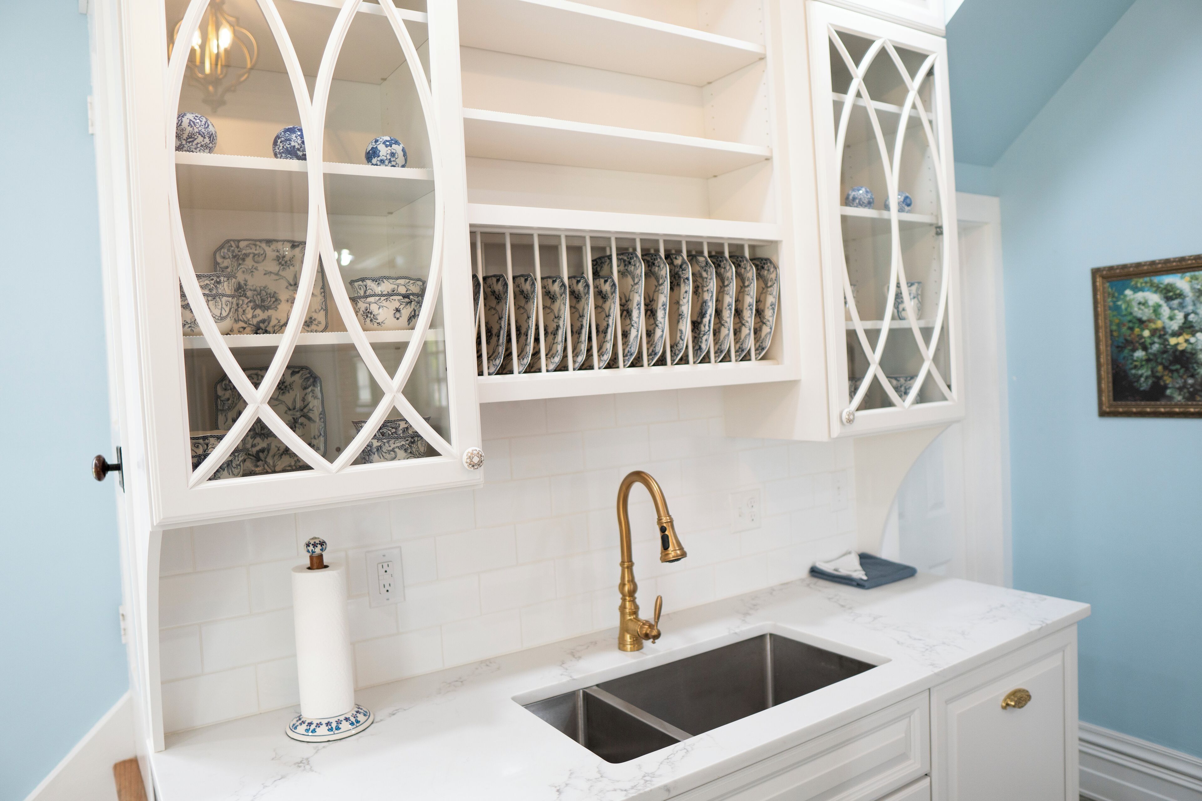 A kitchen with white cabinetry, a marble countertop, a gold faucet, and a dish rack with blue and white plates.