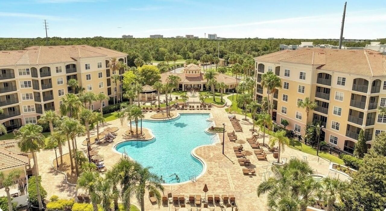 A hotel with a central swimming pool surrounded by lounge areas and palm trees.