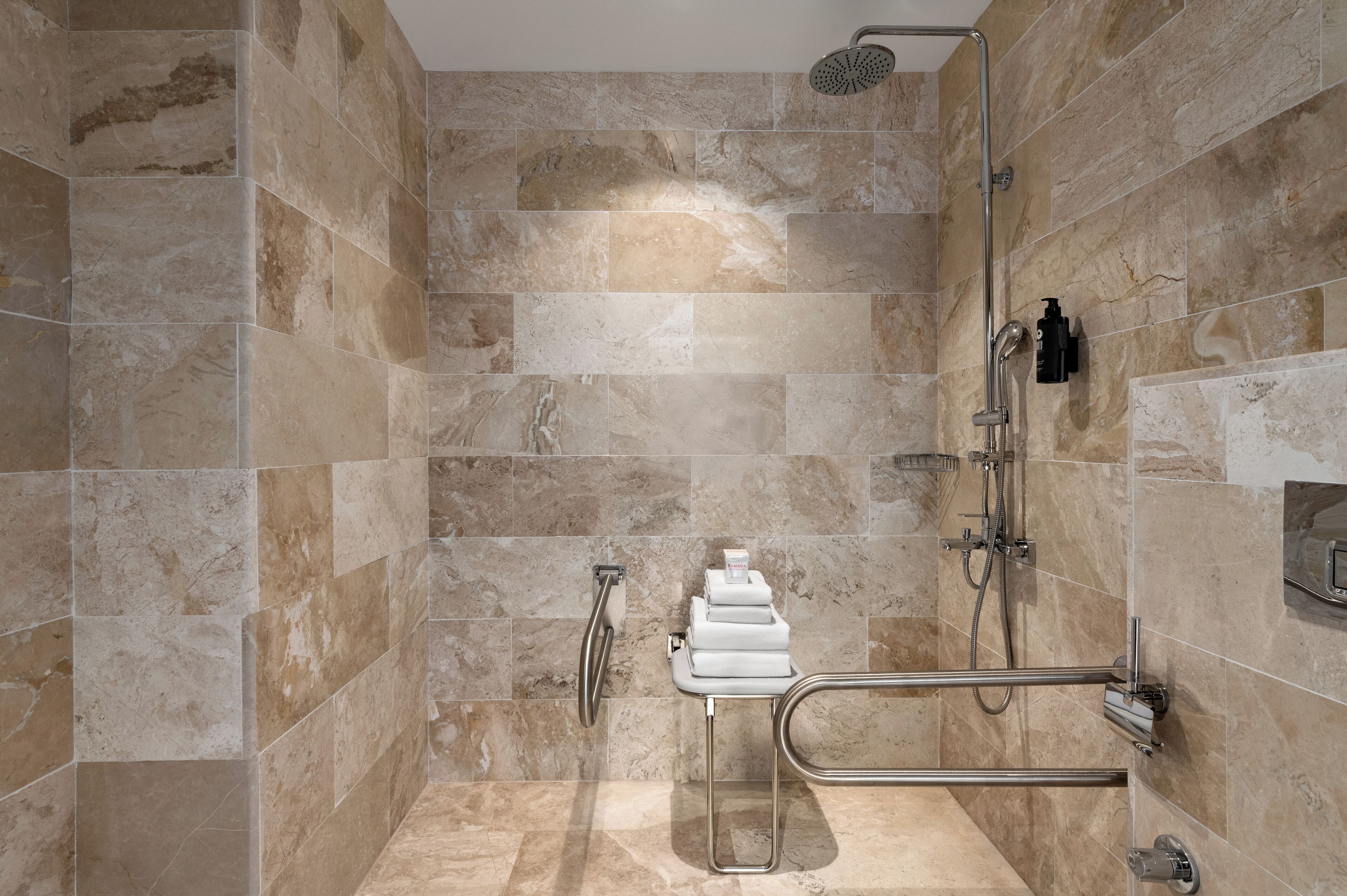 Une salle de bain moderne avec des carreaux de couleur beige, une douche, des toilettes équipées d’une barre d’appui et un distributeur de serviettes.