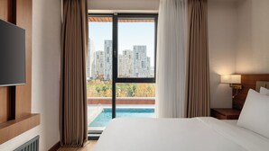 A hotel room with a bed, a TV, a nightstand, a lamp, and a view of a swimming pool and high-rise buildings.