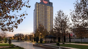 A high-rise hotel with a prominent 'RAMADA RESIDENCE' sign, surrounded by a landscaped park area with a fountain, benches, and a gazebo.