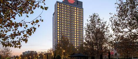 A high-rise hotel with a prominent 'RAMADA RESIDENCE' sign, surrounded by a landscaped park area with a fountain, benches, and a gazebo.