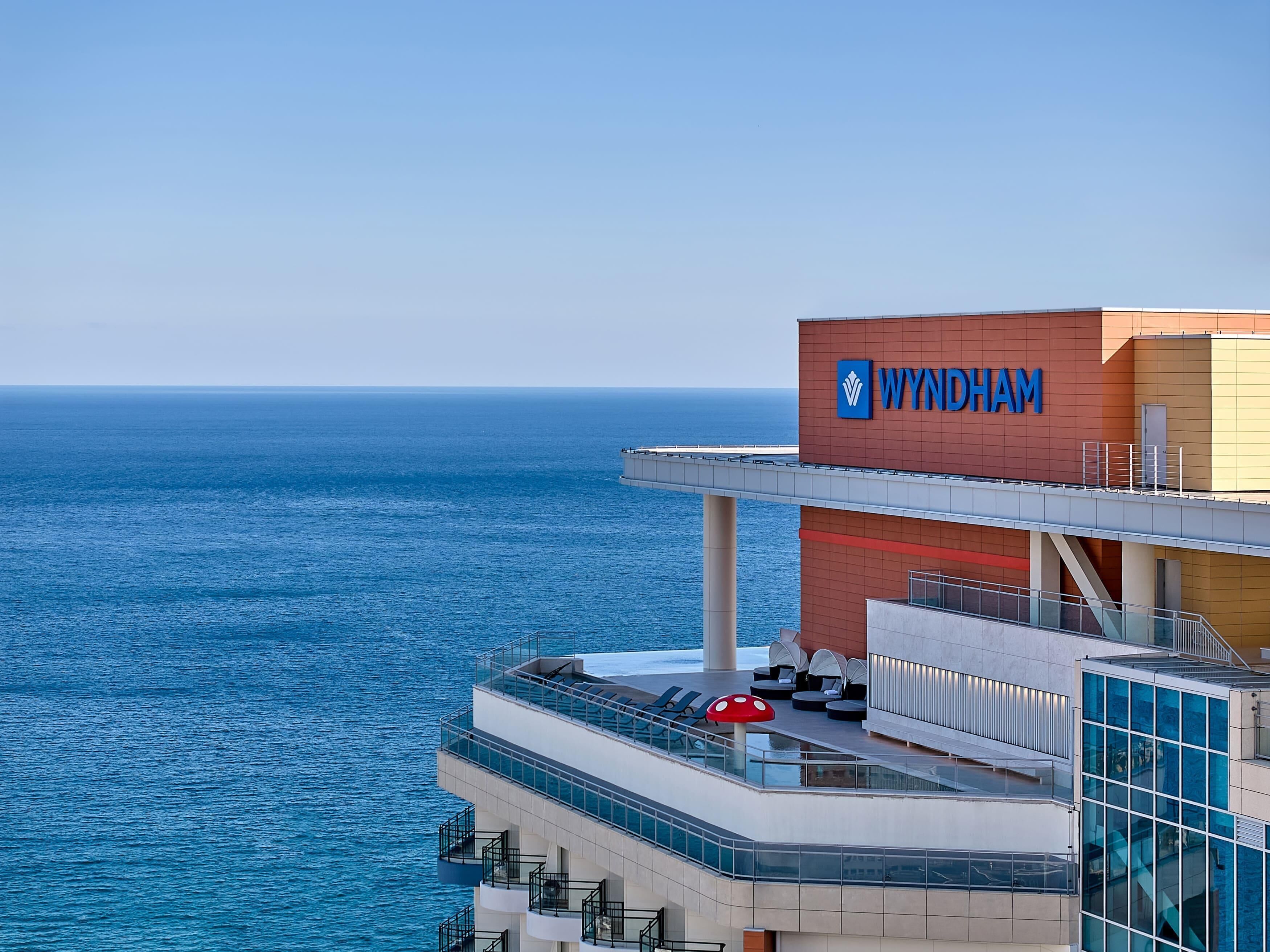 Un hôtel situé en bord de mer, reconnaissable à son grand panneau « WYNDHAM », doté d’un balcon vitré et offrant une vue dégagée sur la mer.
