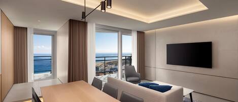 A modern hotel room with a large window offering a view of the ocean, a flat-screen TV mounted on the wall, a wooden dining table, and a sofa with a blue throw pillow.