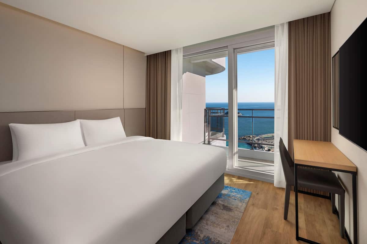 Habitación de hotel con cama, escritorio, silla, televisor y balcón con vista al mar.