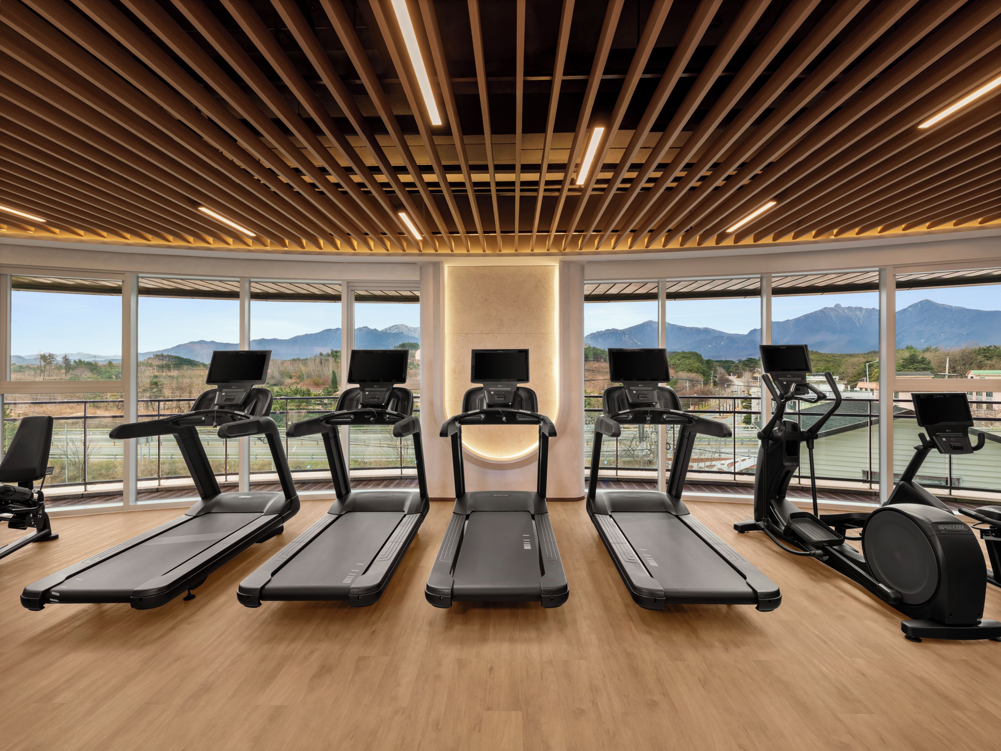 A gym with treadmills and exercise equipment, large windows with a mountain view, and a wooden ceiling design.