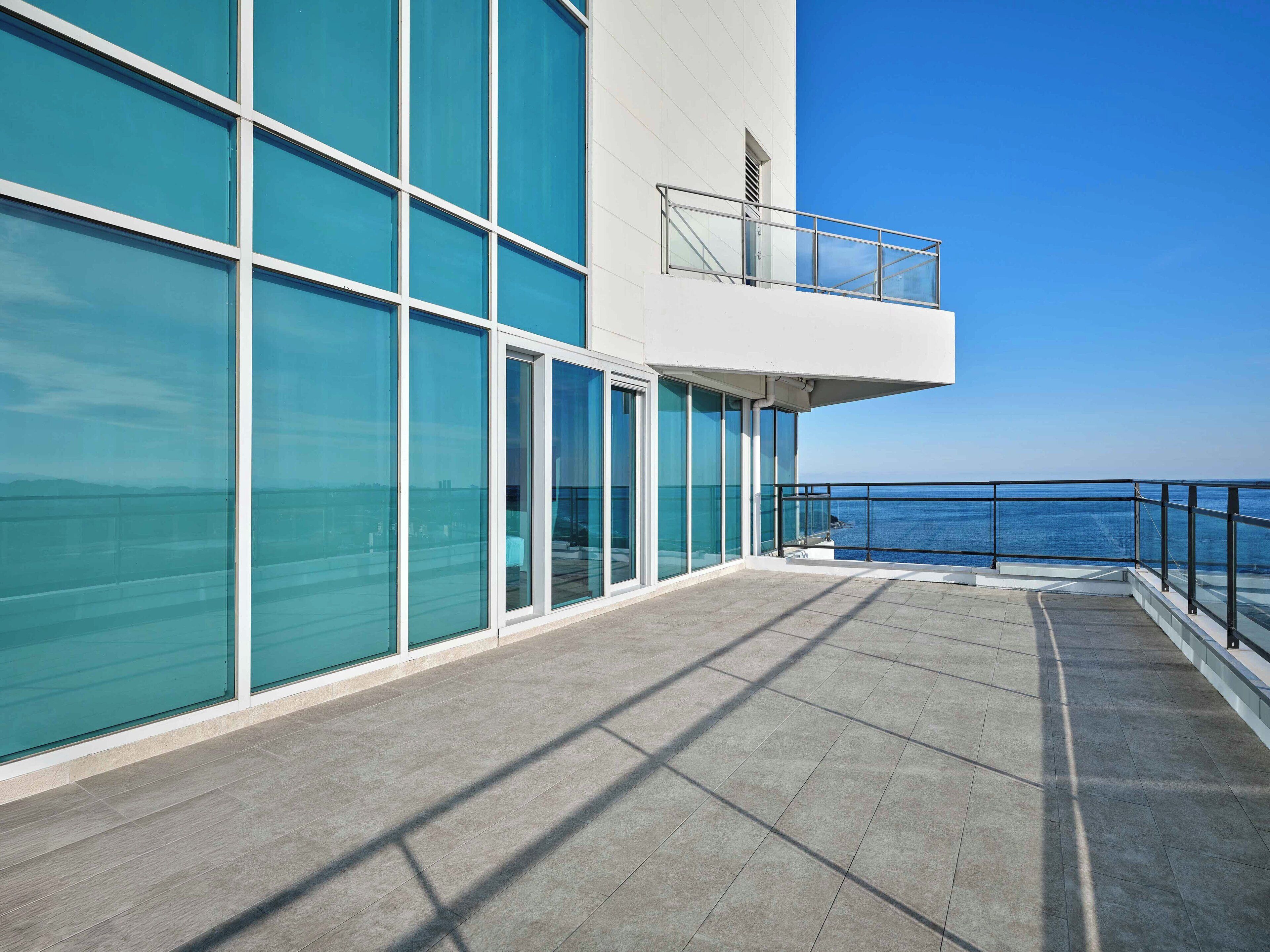 A modern building with large glass windows, a balcony with a metal railing, and a view of the sea.