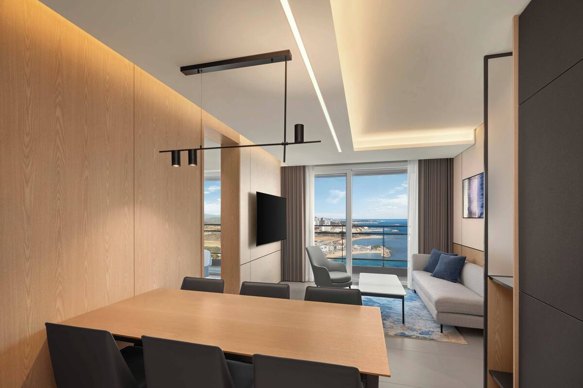 Habitación de hotel moderna con comedor, sofá, televisor y ventana grande con vista al mar.