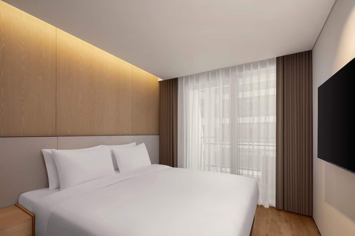 Una habitación de hotel moderna con una cama grande, cabecera de madera, un televisor de pantalla plana y cortinas translúcidas.