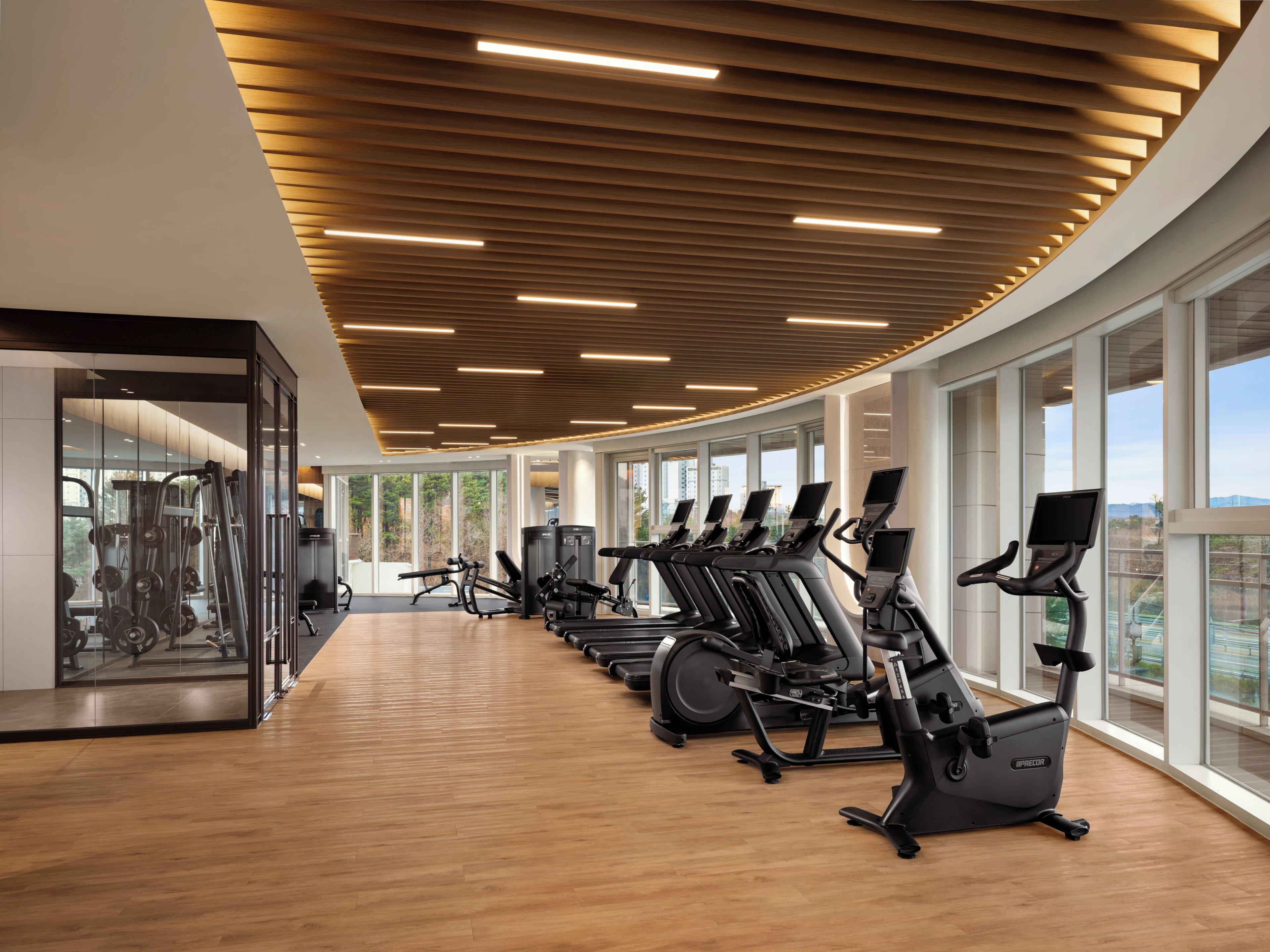 Academia com equipamentos de ginástica, janelas amplas e teto de madeira.