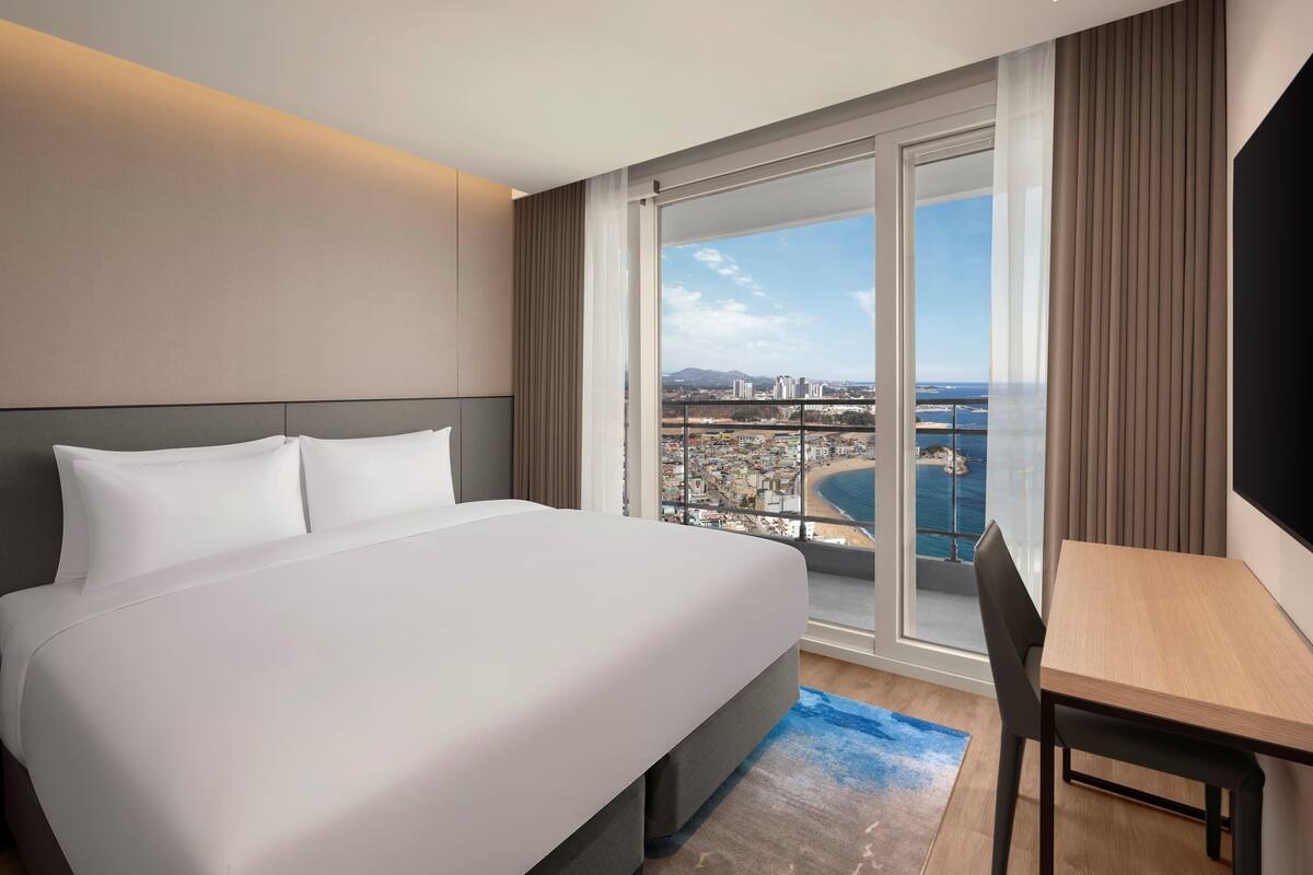 Habitación de hotel con una cama grande, un escritorio y vistas a la ciudad y a la costa.