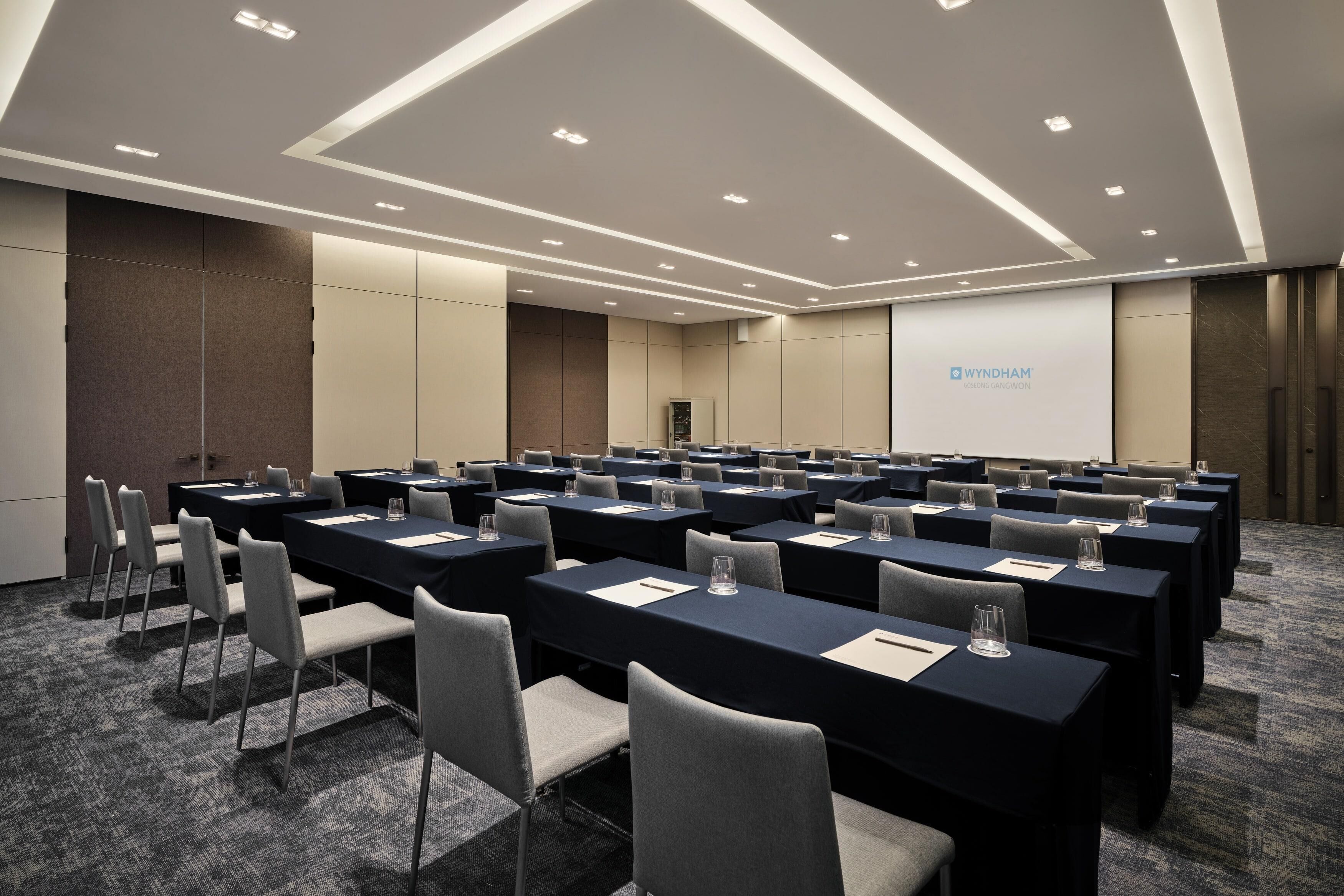 A conference room with rows of tables and chairs, a projection screen, and a podium.