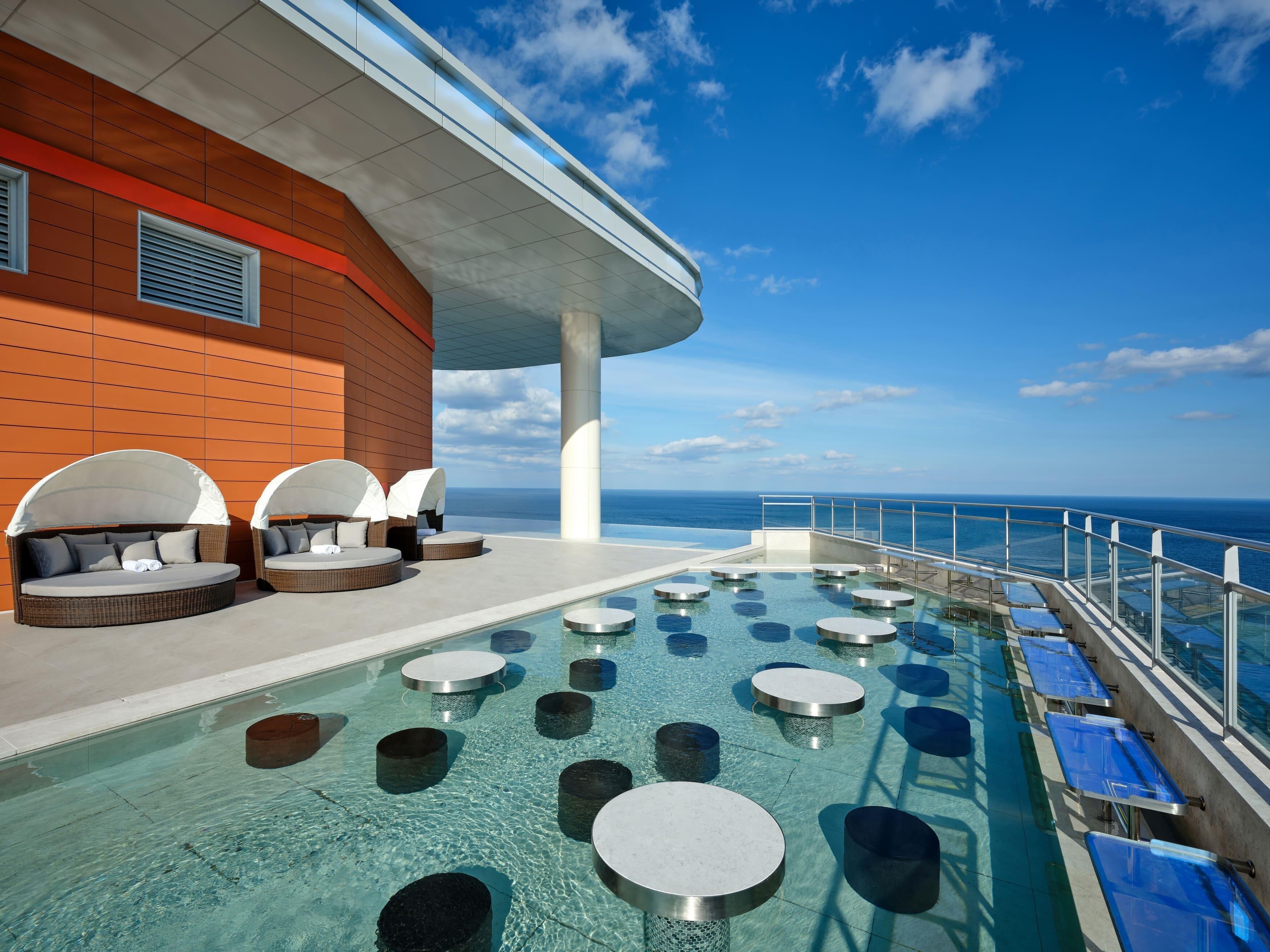 A pool area with lounge chairs and tables, a clear view of the ocean, and a modern building structure.