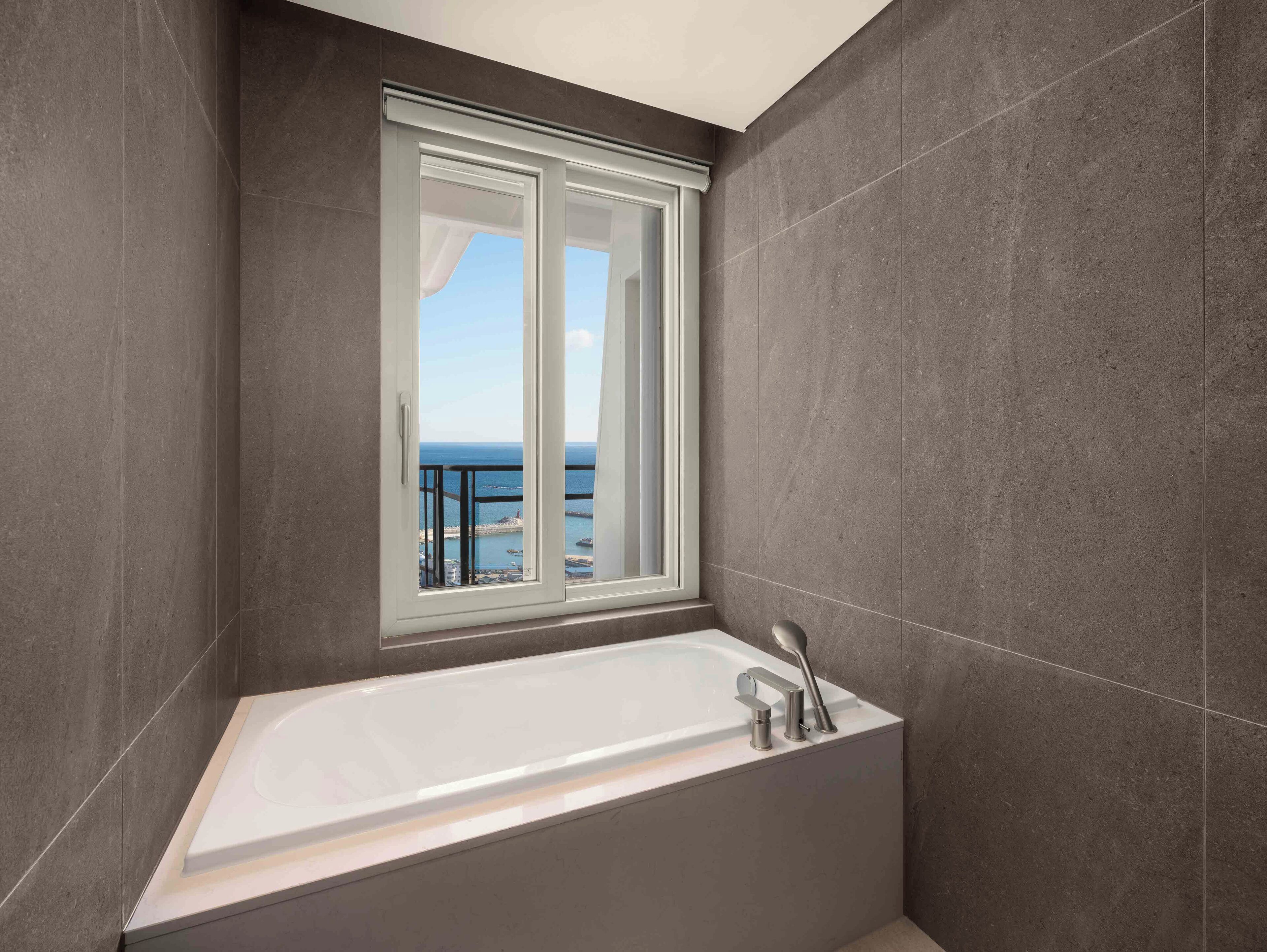 A modern bathroom with a bathtub, a window with a view of the sea, and grey tiled walls.