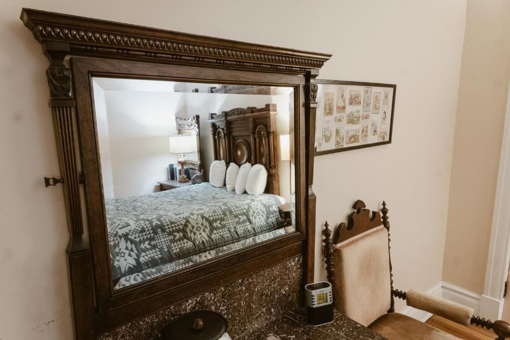 A bedroom with a large mirror, a bed with patterned bedding, a wooden headboard, a small table, and a framed picture on the wall.