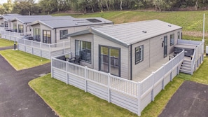 A row of modern, single-story mobile homes with balconies, a covered outdoor seating area, and a grassy landscape in the background. - Cedar Lodge 2 Bedrooms With hot tub (Northwich)