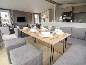 A modern living room with a dining area set for six, grey sofas, a flat-screen TV, and a kitchen with a microwave. - Cedar Lodge 2 Bedrooms With hot tub (Northwich)
