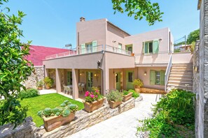 A two-story building with a stone facade, a balcony, and a well-maintained garden with potted plants. - Villa Verde Petra - Three Bedroom Villa, Sleeps 6 (Rethymno)