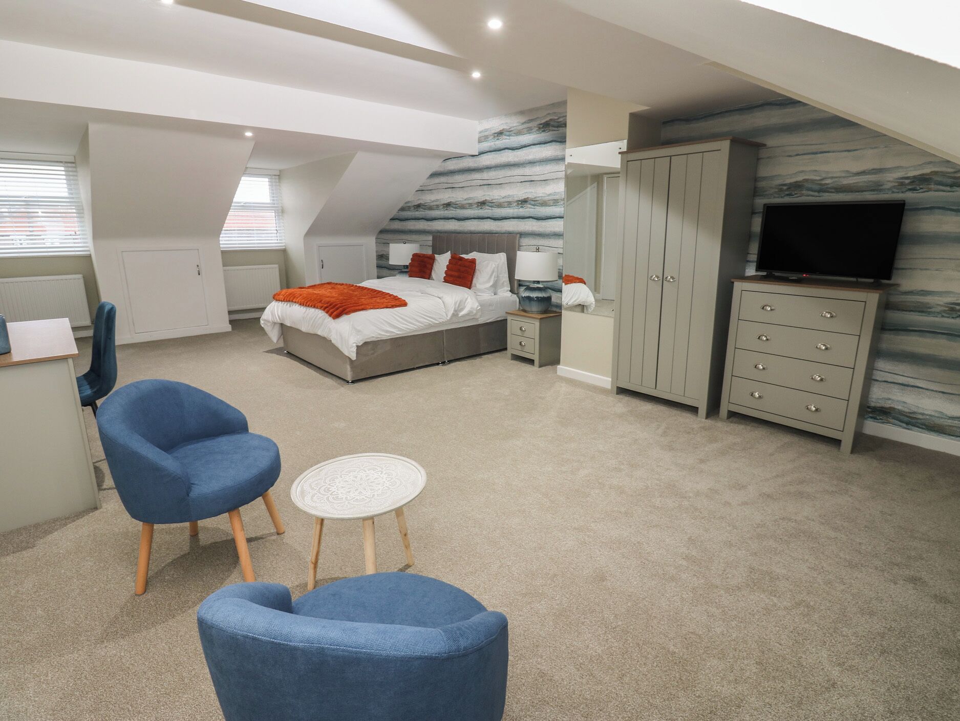 A bedroom with a bed, two blue chairs, a small white table, a TV, and a wardrobe.