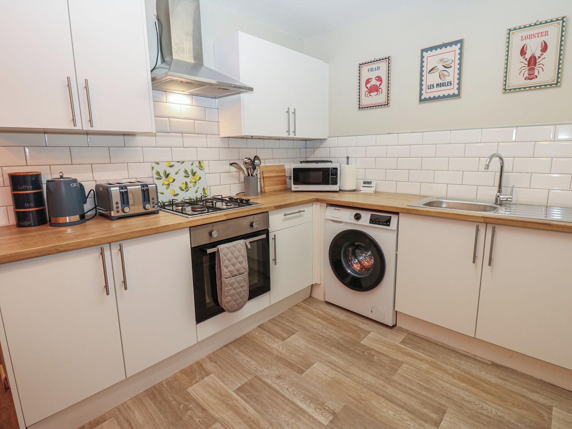 A modern kitchen with white cabinets, a microwave, a toaster, a kettle, a gas stove, a washing machine, and a sink.