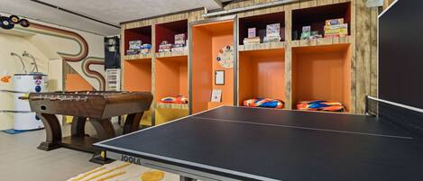 A room with a foosball table, ping pong table, and shelves with books and games.