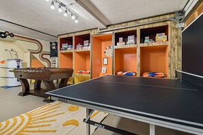 A room with a foosball table, ping pong table, and shelves with books and games.
