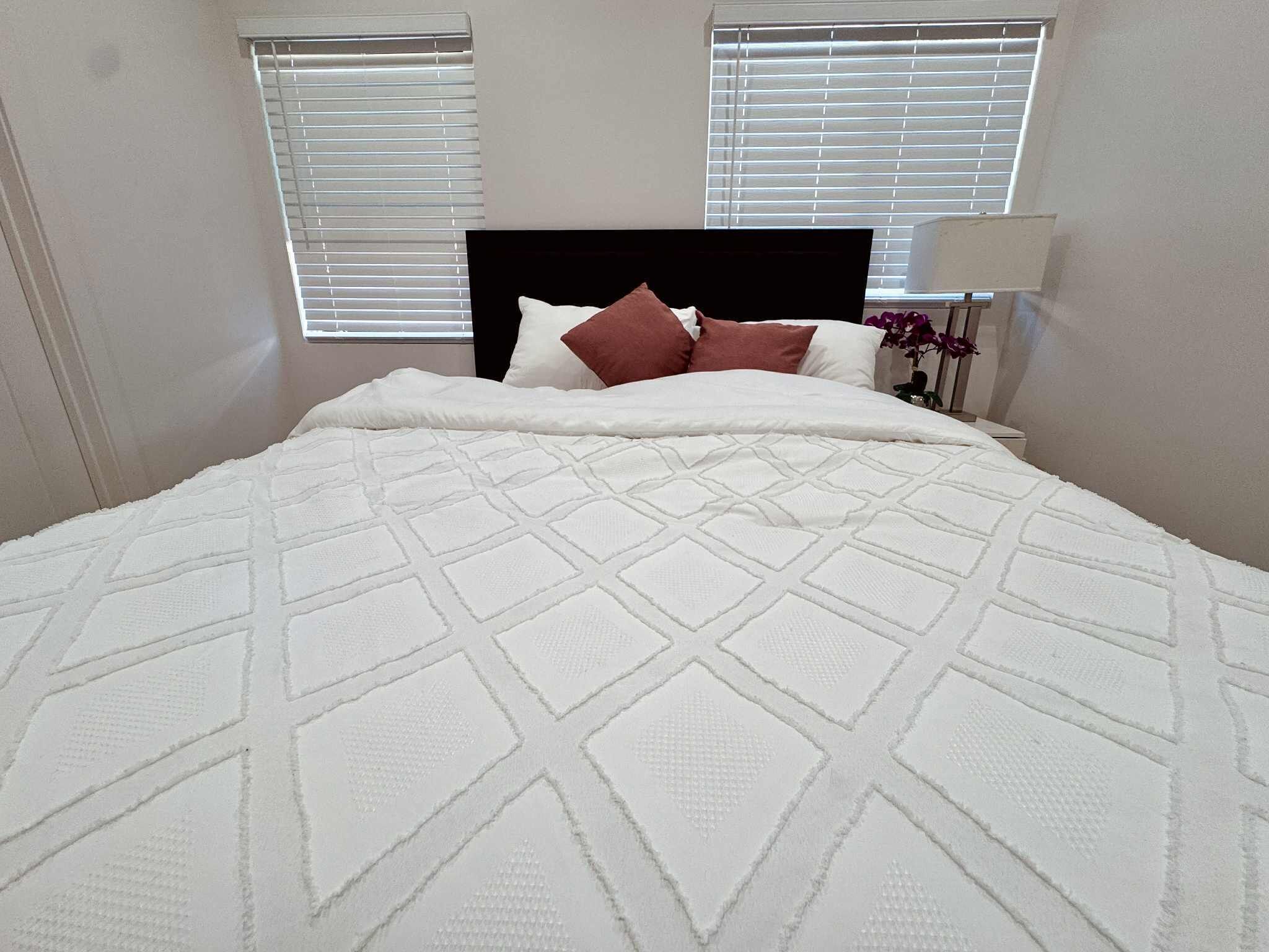 A neatly made bed with a white quilt and two pillows, a bedside lamp, and a window with blinds.