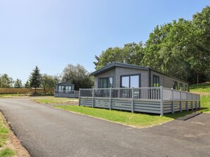 Ein modernes Mobilheim mit umzäunter Terrasse, umgeben von viel Grün und blauem Himmel.
