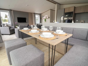 Un salon moderne avec un coin repas aménagé pour six personnes, des canapés gris, un téléviseur à écran plat et une cuisine équipée d’un four à micro-ondes.