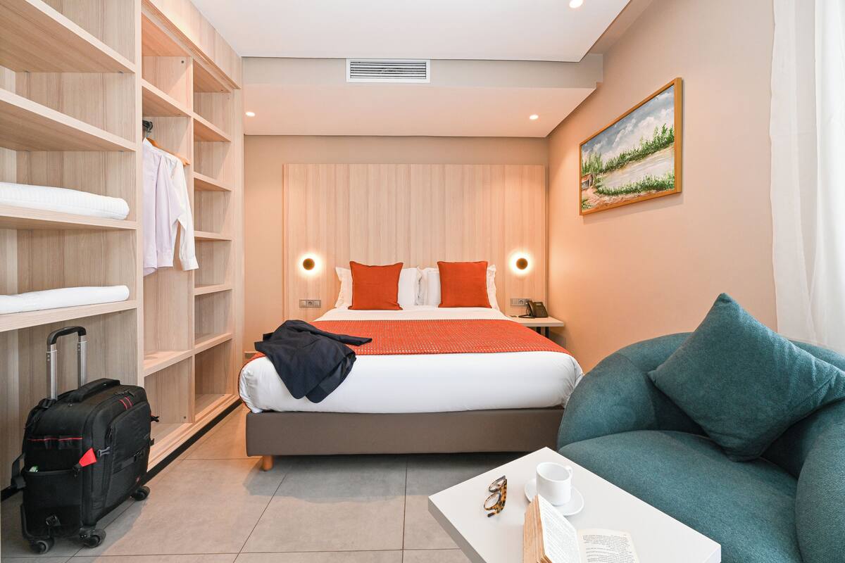 A hotel room with a bed, a sofa, a desk with a cup and book, a suitcase, and a wardrobe.